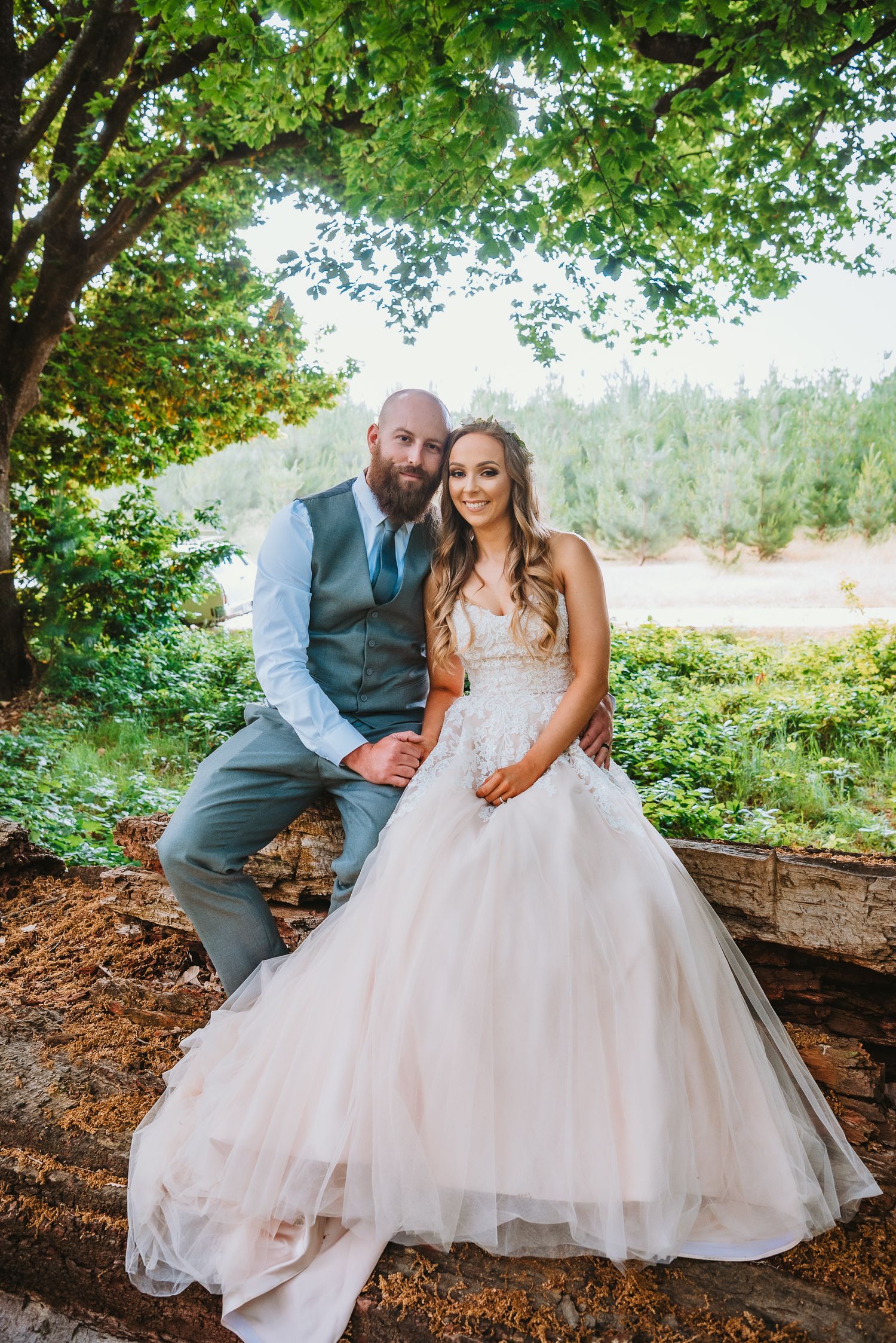 A wedding couple sits together on a rustic log beneath lush green trees at an outdoor forest venue.
