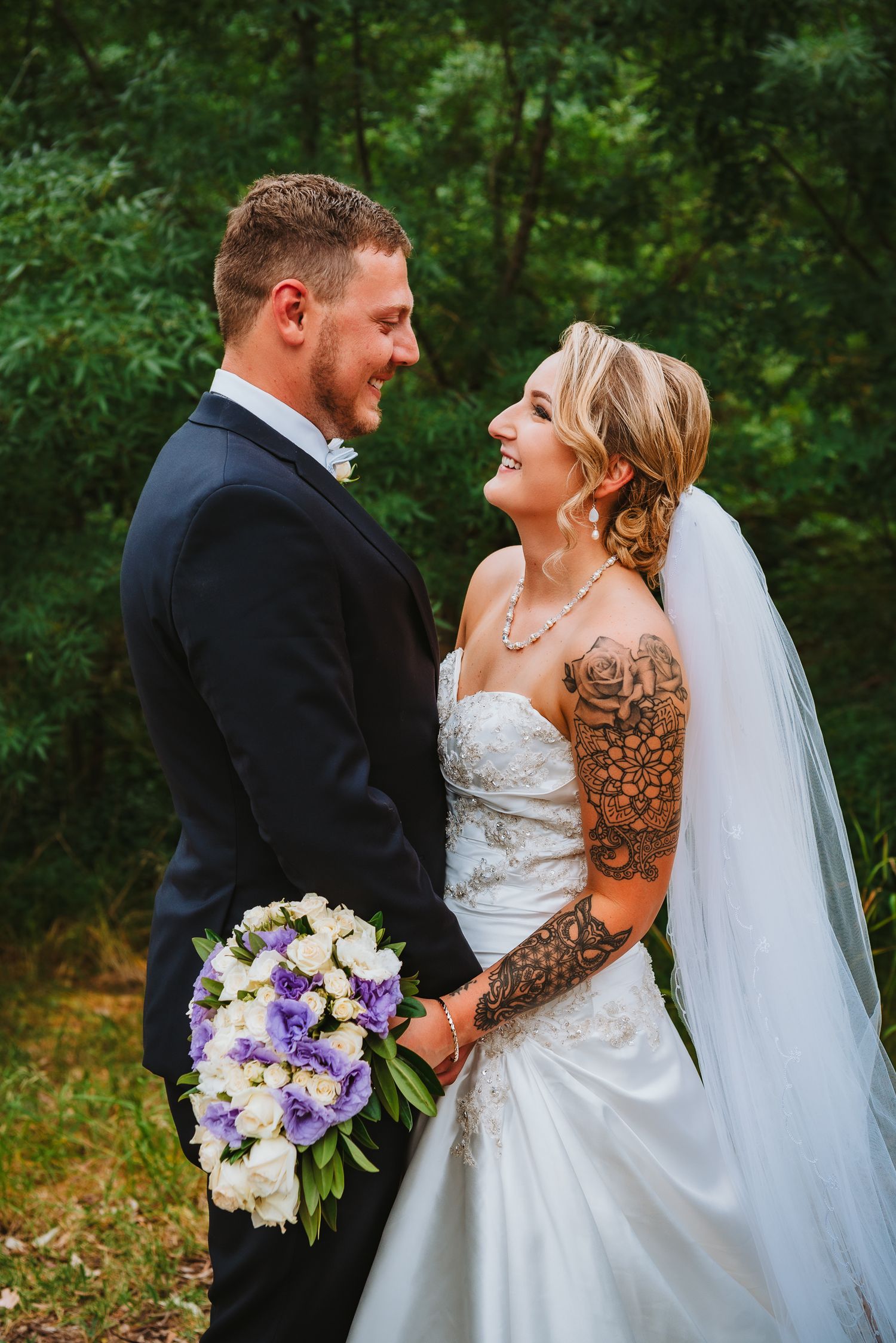 A bride in a white dress with sleeve tattoos holds a purple and white bouquet while laughing with her groom in a forest.