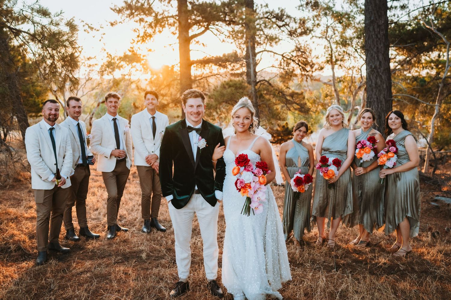 Wedding party poses in rustic forest setting at sunset with bridesmaids in sage dresses and groomsmen in light blue attire.