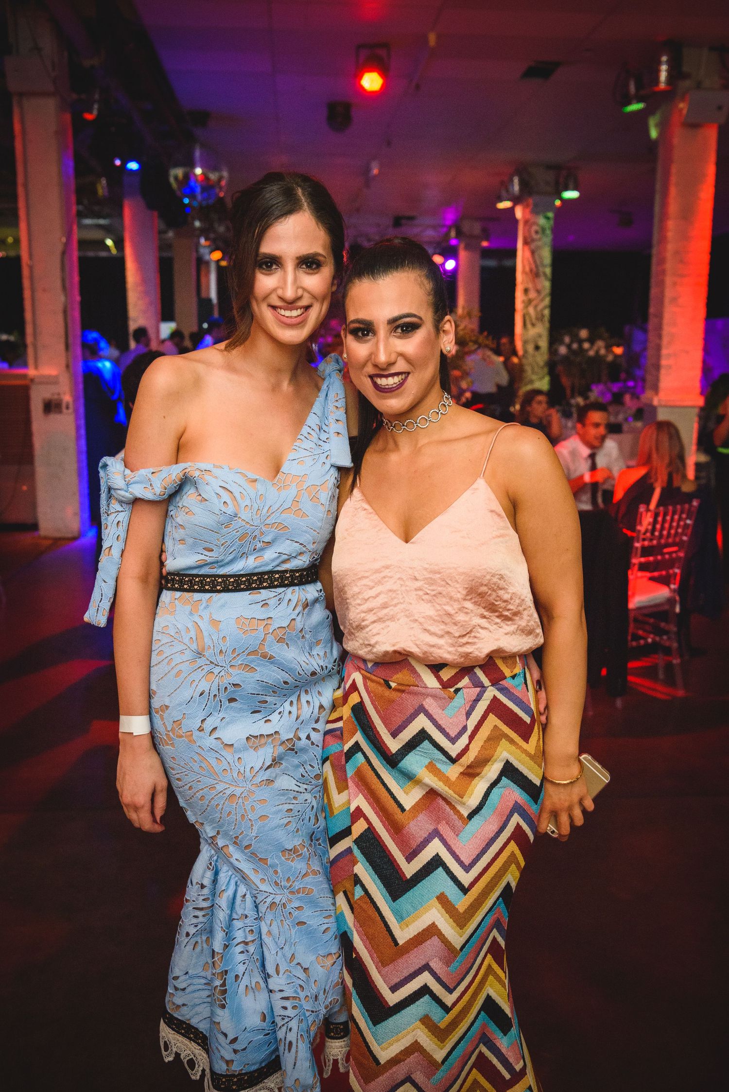 Two women pose at a party venue wearing evening dresses - one in blue lace and one in a colorful chevron pattern skirt.