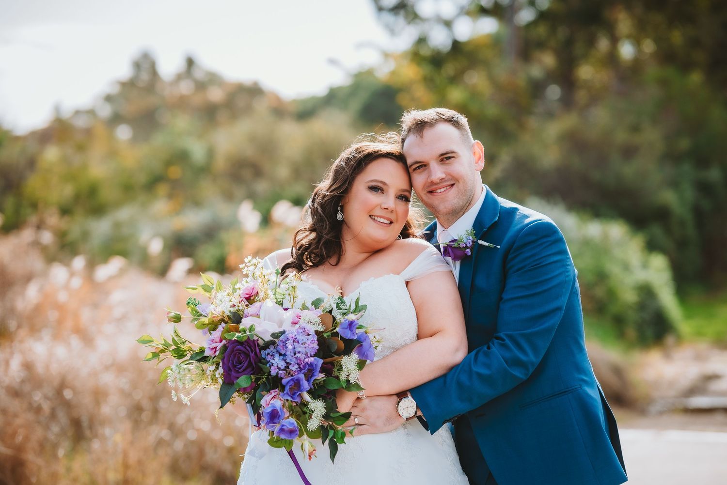 A joyful couple in wedding attire embraces outdoors while holding a colorful purple and white floral bouquet.