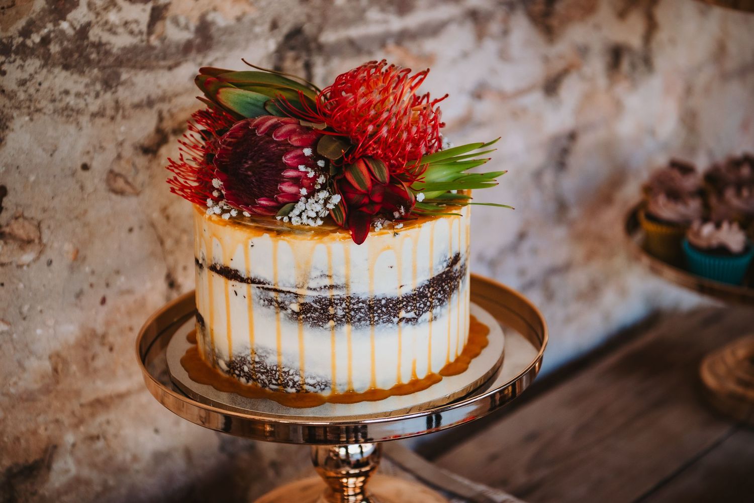 A small single-tiered wedding cake decorated with red protea flowers and berries on a gold cake stand.
