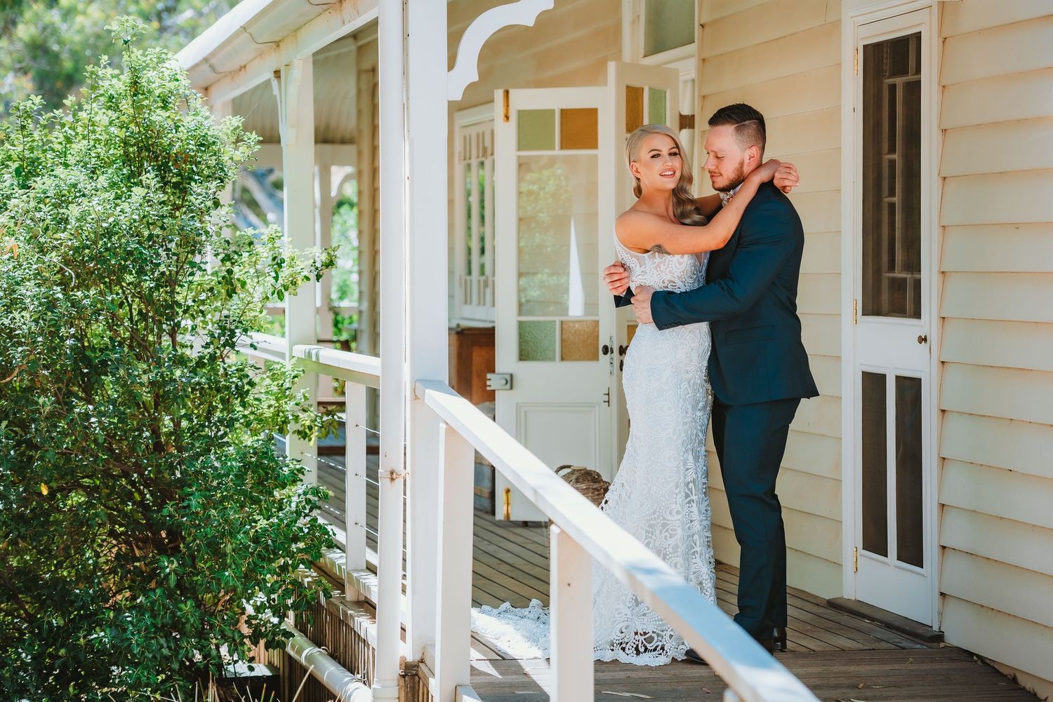 A couple embraces on the porch of a white farmhouse with greenery surrounding them.