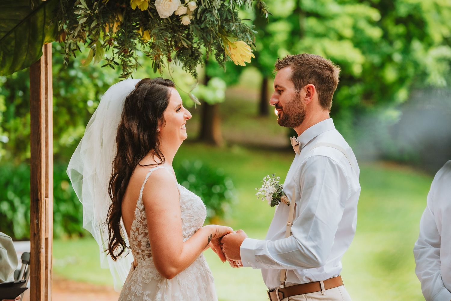 A couple shares a joyful moment during their outdoor wedding ceremony under a floral arch in a green garden setting.