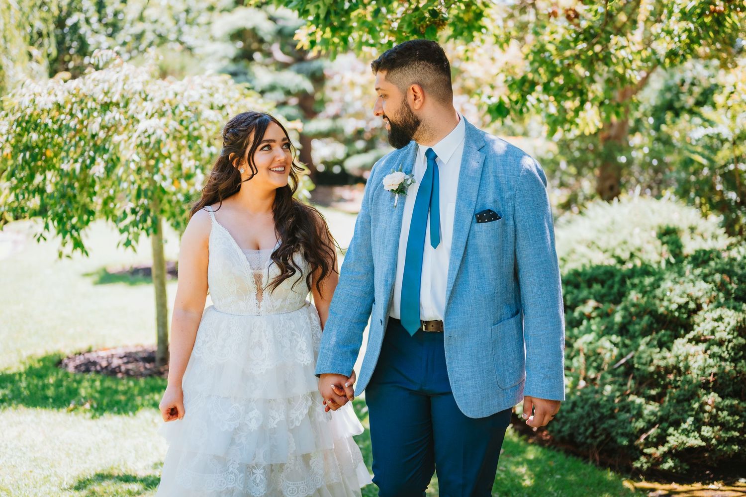 A couple shares a joyful moment together in a garden setting, wearing formal wedding attire on a sunny day.