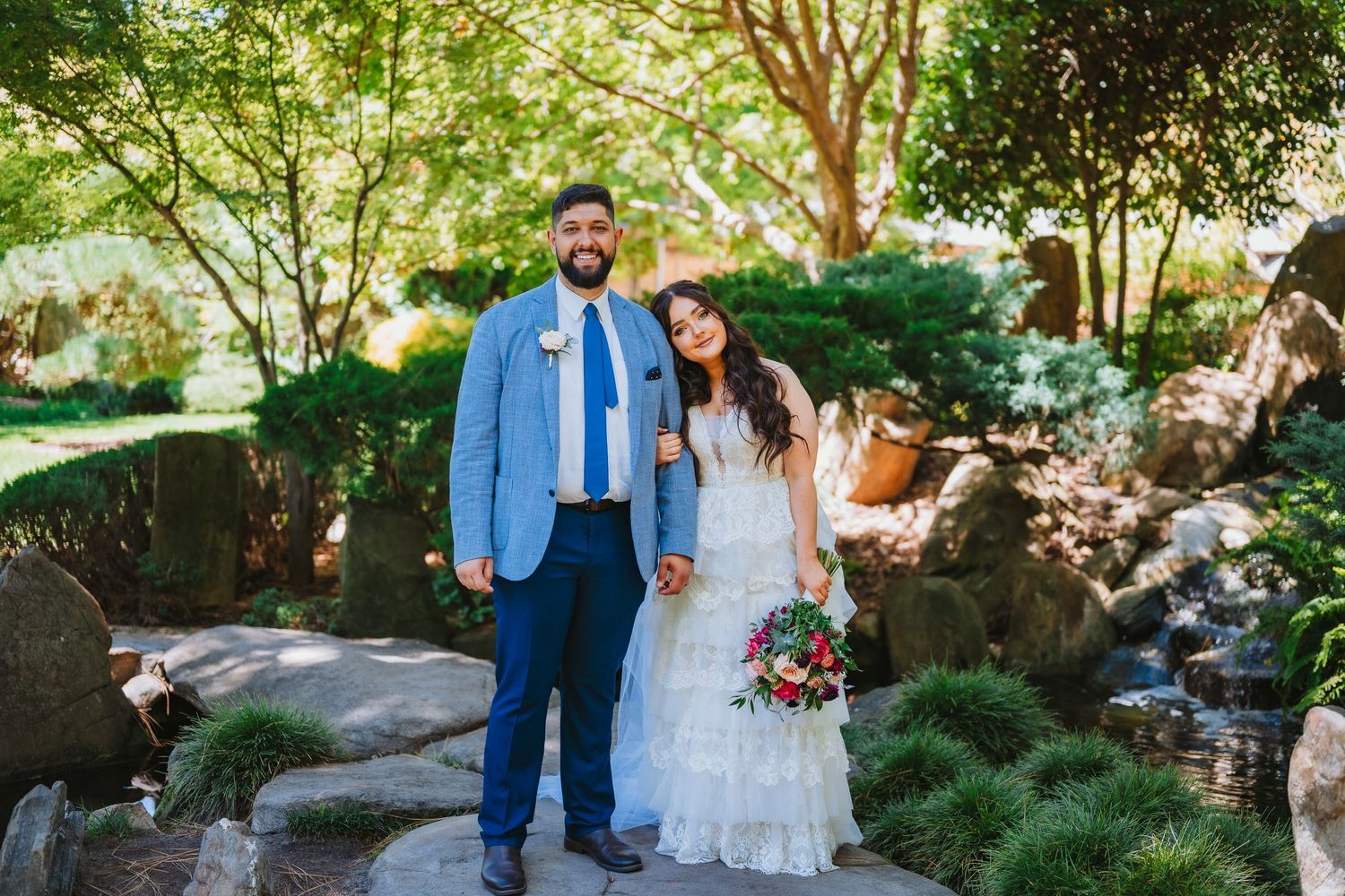 A couple in wedding attire stands together in a garden setting with lush greenery and natural stone features.