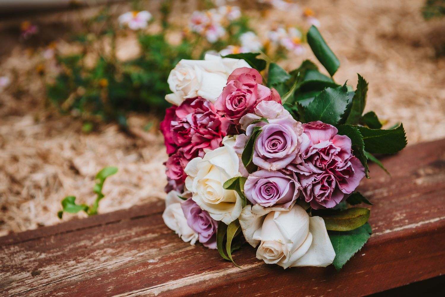 A romantic wedding bouquet featuring pink, purple and white roses resting on a rustic wooden bench.