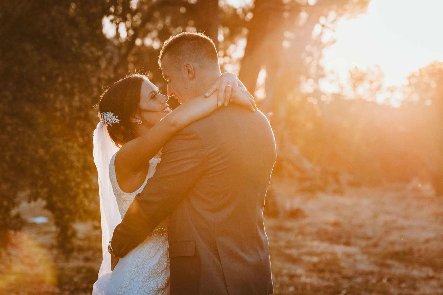 Silhouetted couple embracing during golden hour sunset with sunlight streaming through trees in the background.