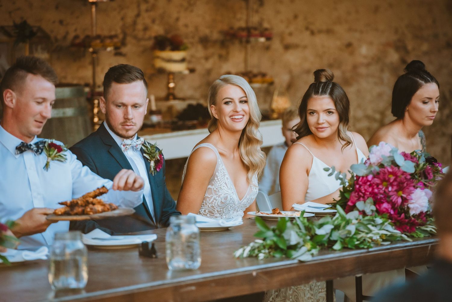 Wedding party sits together at rustic reception table with colorful floral arrangements and table settings.