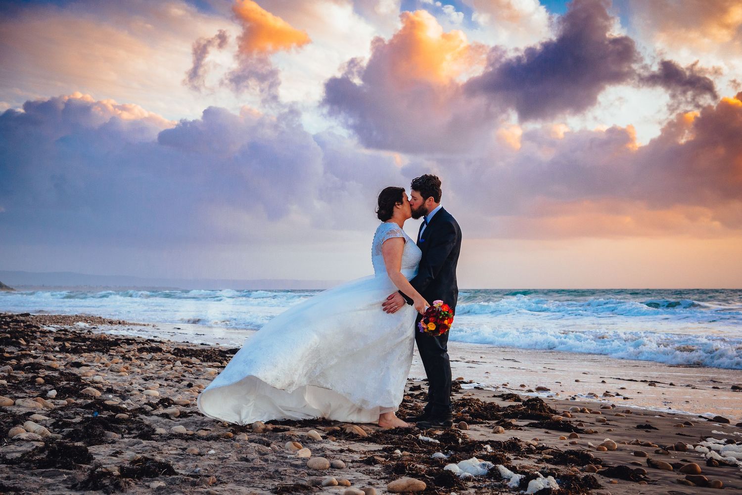 A romantic wedding couple embraces on a beach at sunset with dramatic orange clouds and crashing waves in the background.