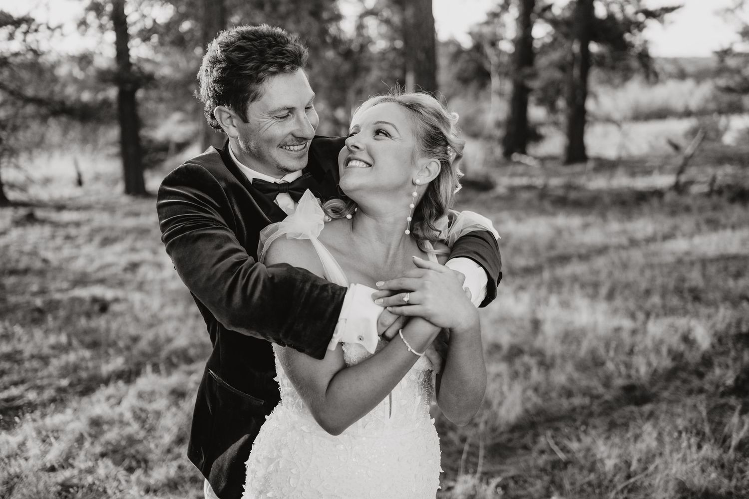A black and white portrait of a couple embracing and laughing outdoors in a wooded area.