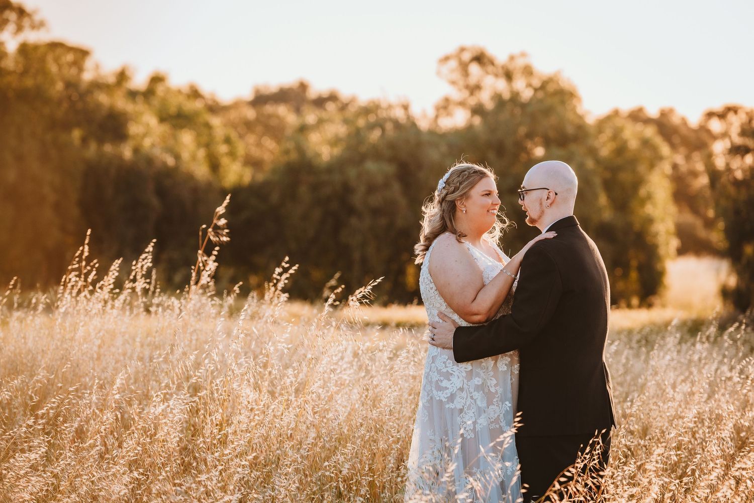 Couple embraces in a sunlit field of golden grass during sunset with trees in background.
