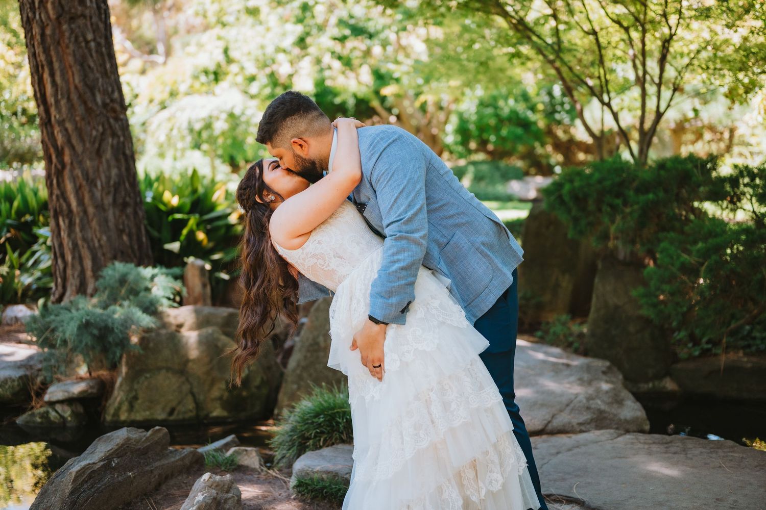 A couple in wedding attire embraces while dipping in a romantic pose surrounded by lush garden scenery.