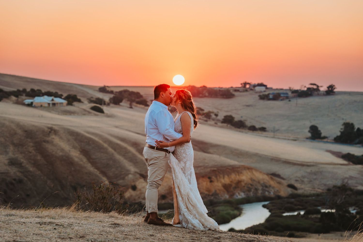 A couple embraces on a hilltop during sunset with rolling golden hills in the background.