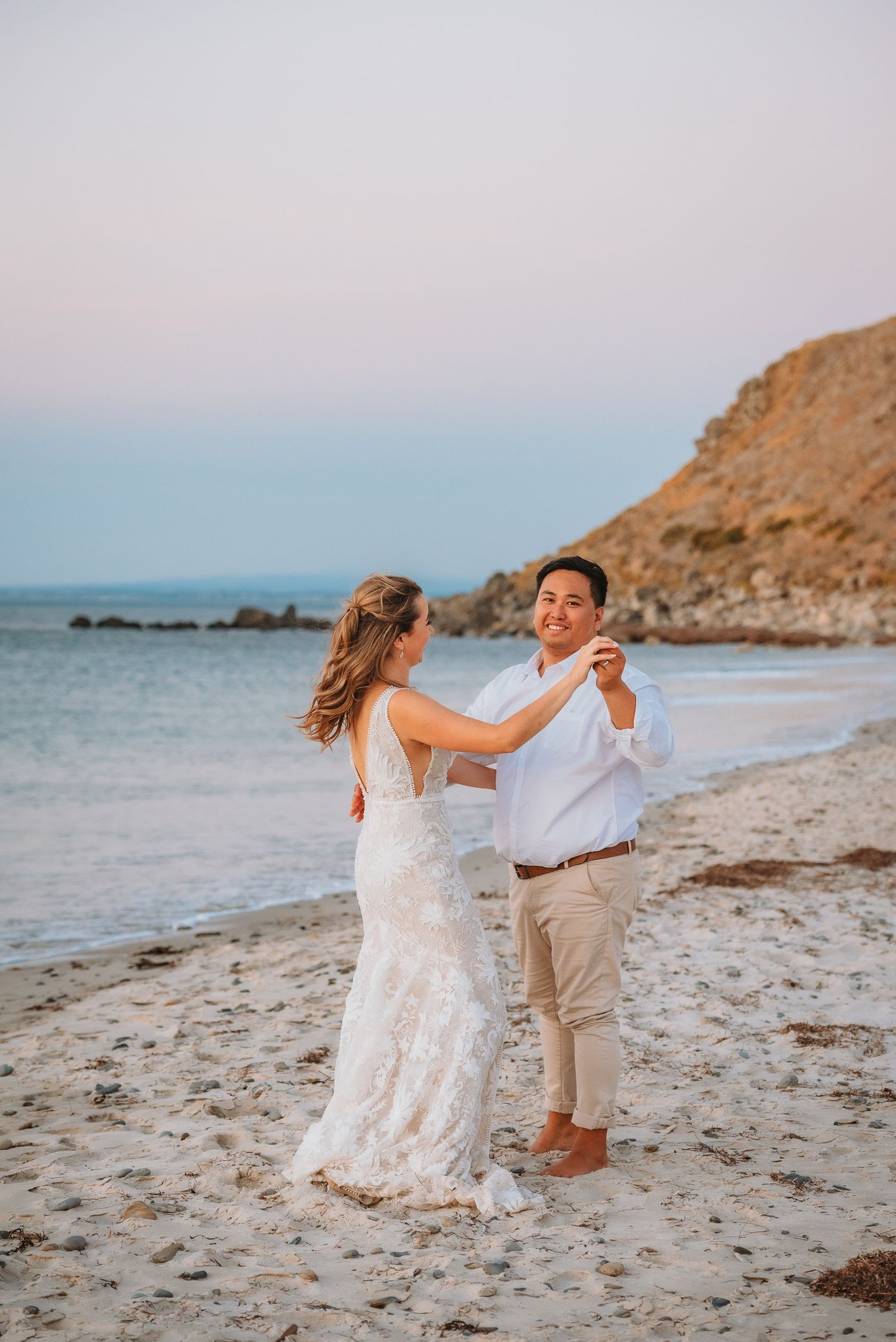 A couple dancing together on a scenic beach at sunset with cliffs and ocean in the background.
