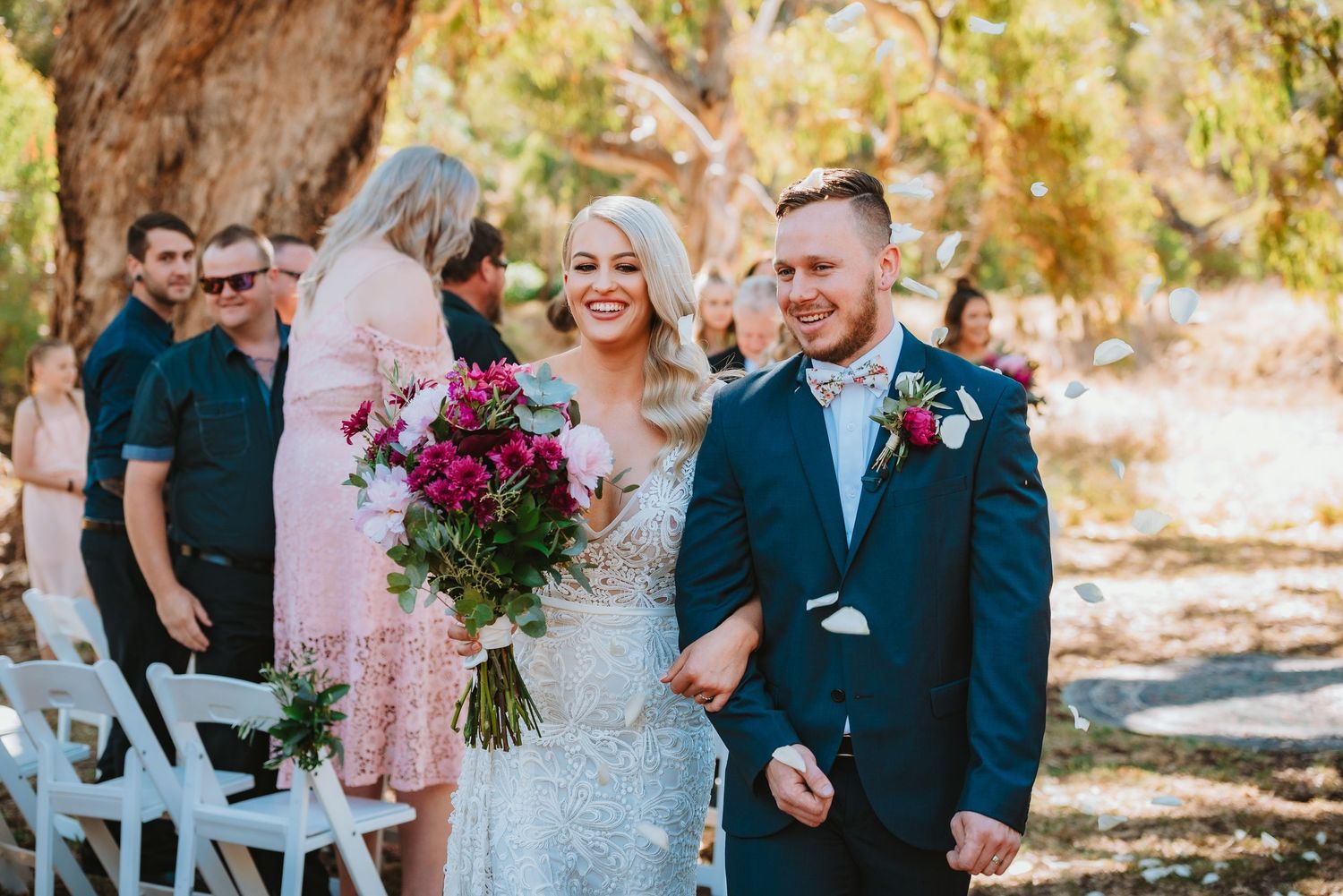 A wedding ceremony takes place outdoors in a wooded setting with white chairs and beautiful burgundy floral bouquets.