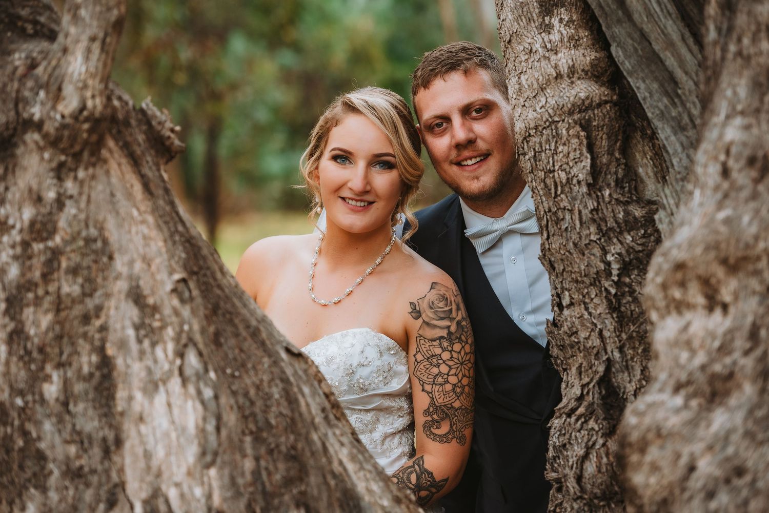 A newly married couple poses between large tree trunks in a natural outdoor setting, with the bride wearing a white dress.