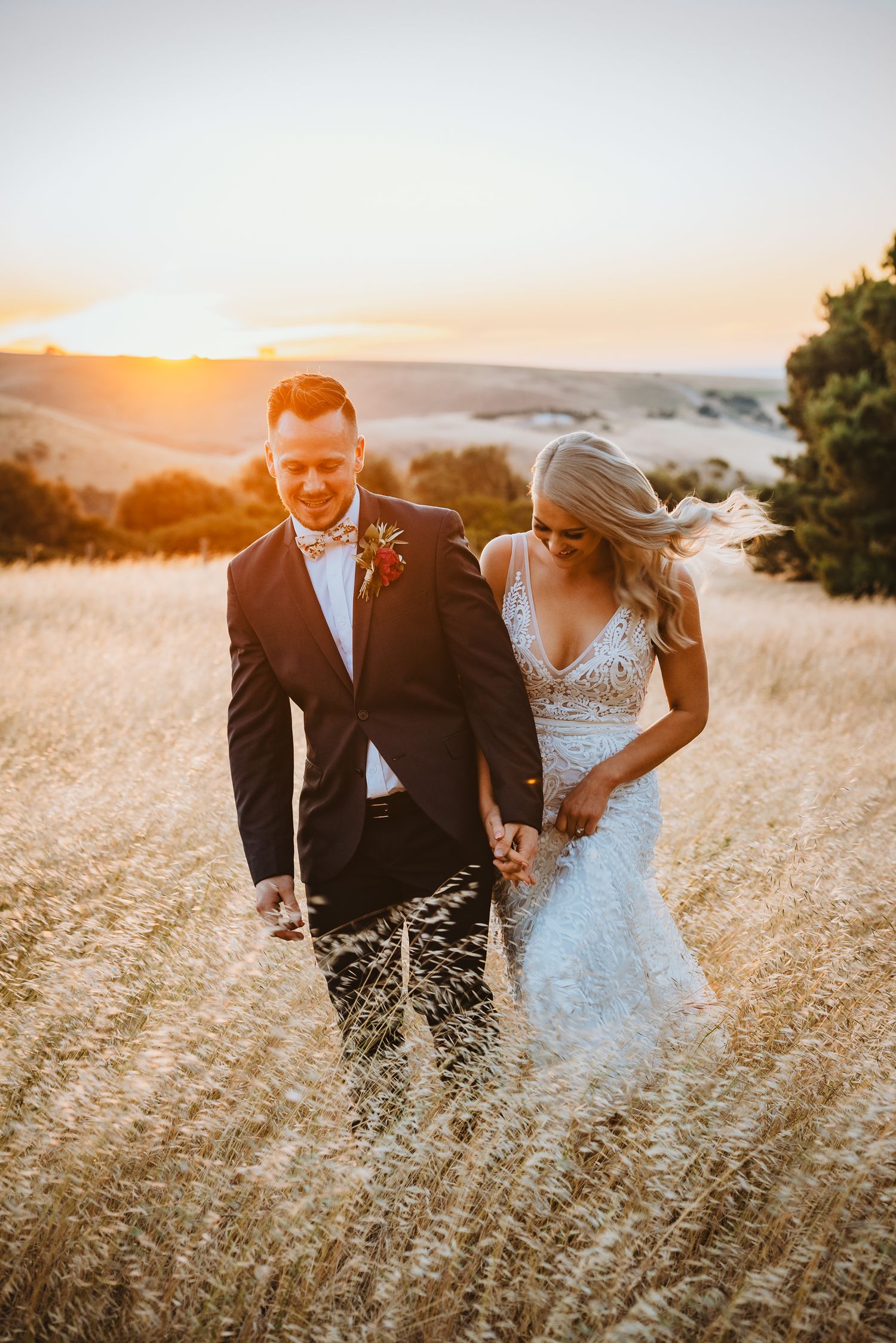 Golden sunset light illuminates couple walking through tall grass field in formal wedding attire during golden hour.