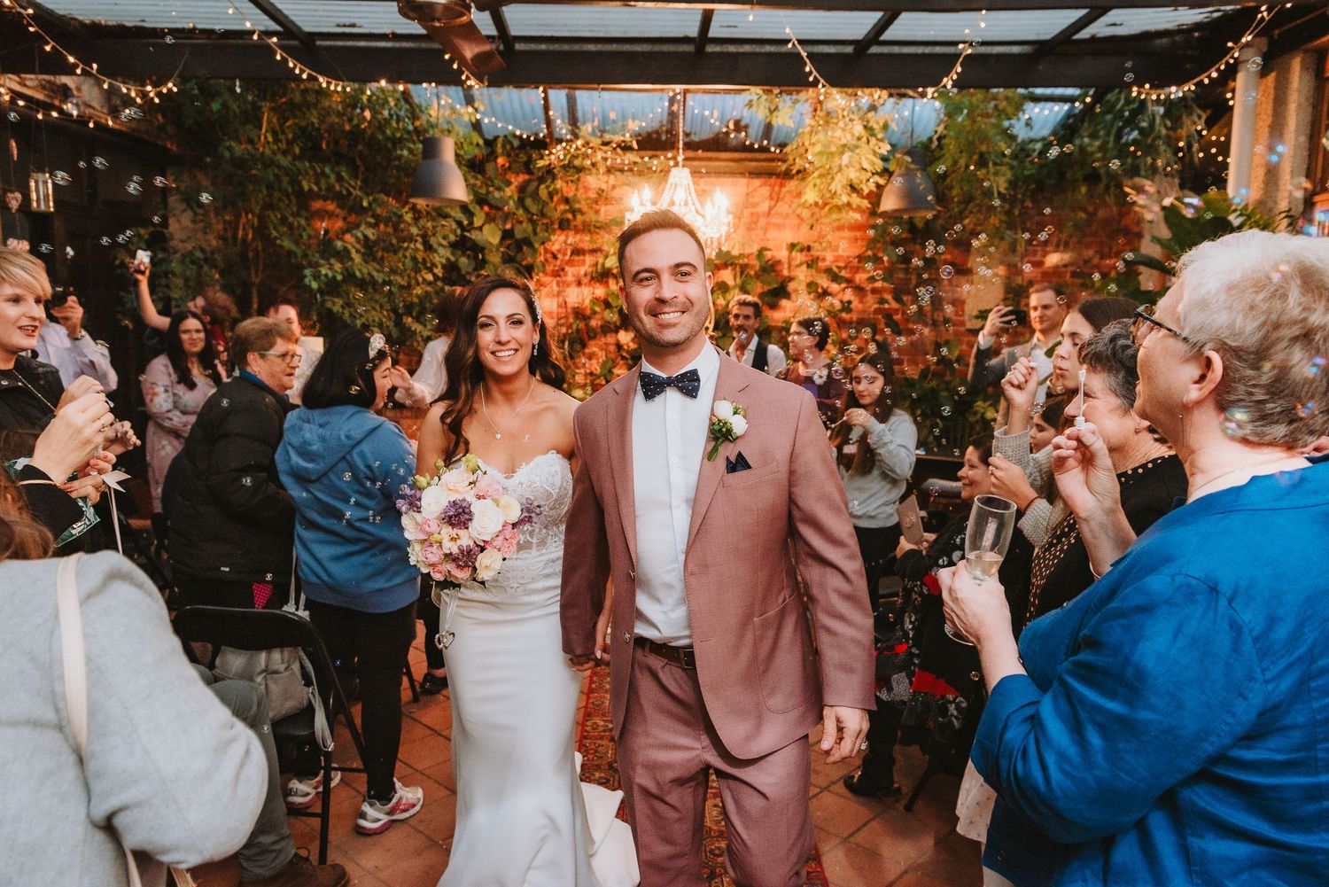 A celebratory wedding reception scene with string lights, greenery decor and guests cheering in an indoor venue.