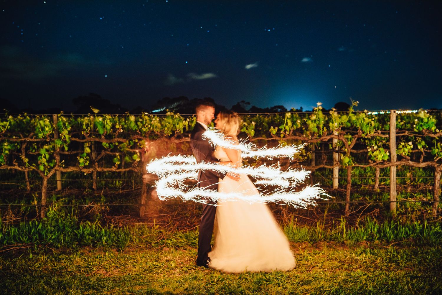 Light painting creates a spinning heart effect against a starry night sky in a vineyard setting.
