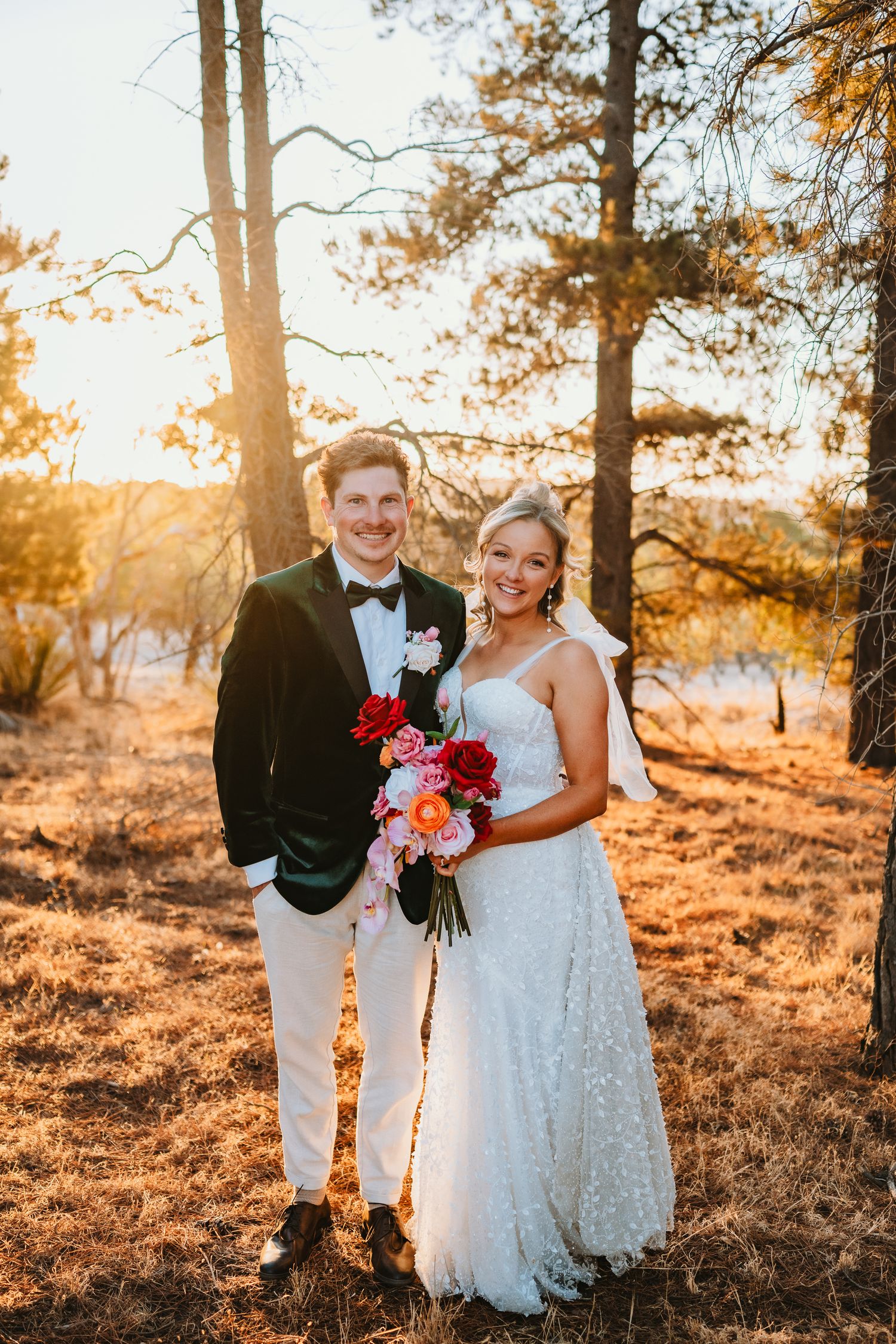 A wedding couple stands in a forest at golden hour with sunlight filtering through pine trees in warm autumn colors.