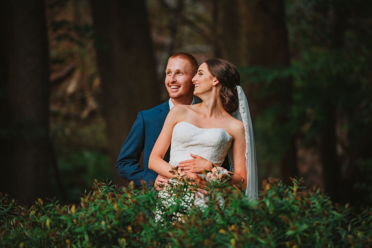 A happy newlywed couple embraces in a forest setting with lush greenery during golden hour photography.