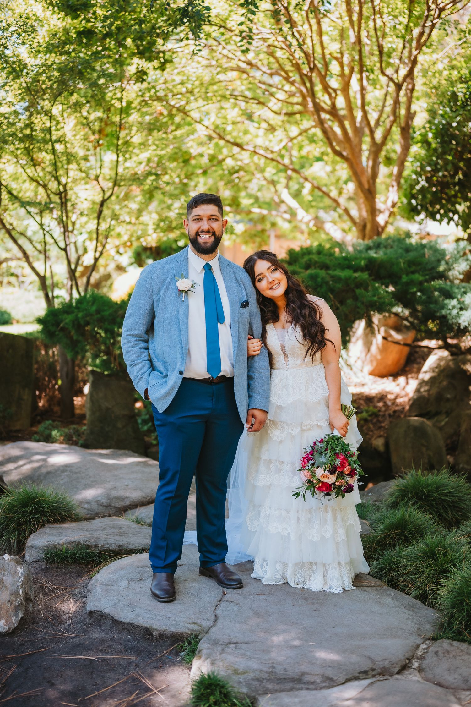 A happy couple stands together outdoors in a garden setting on their wedding day, surrounded by lush greenery.