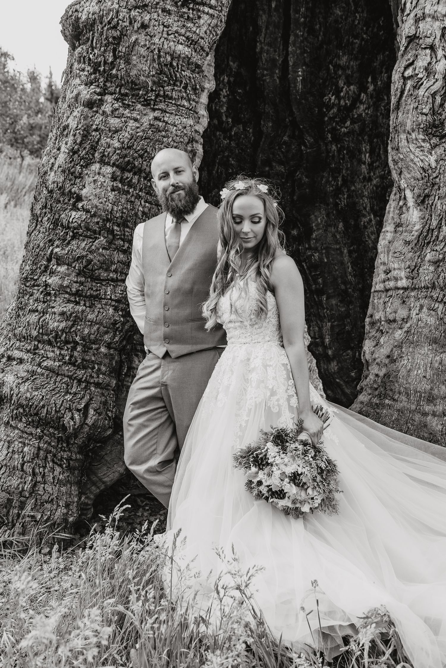 A bride and groom pose against large palm tree trunks in a black and white wedding portrait.