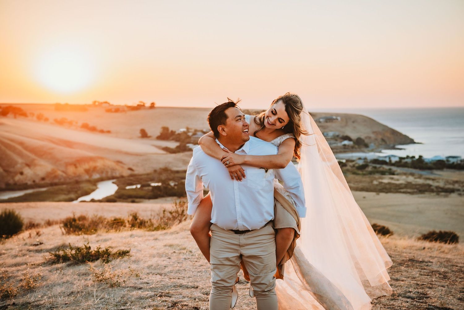 A couple embraces on a coastal hilltop during golden hour sunset with ocean and cliffs in the background.