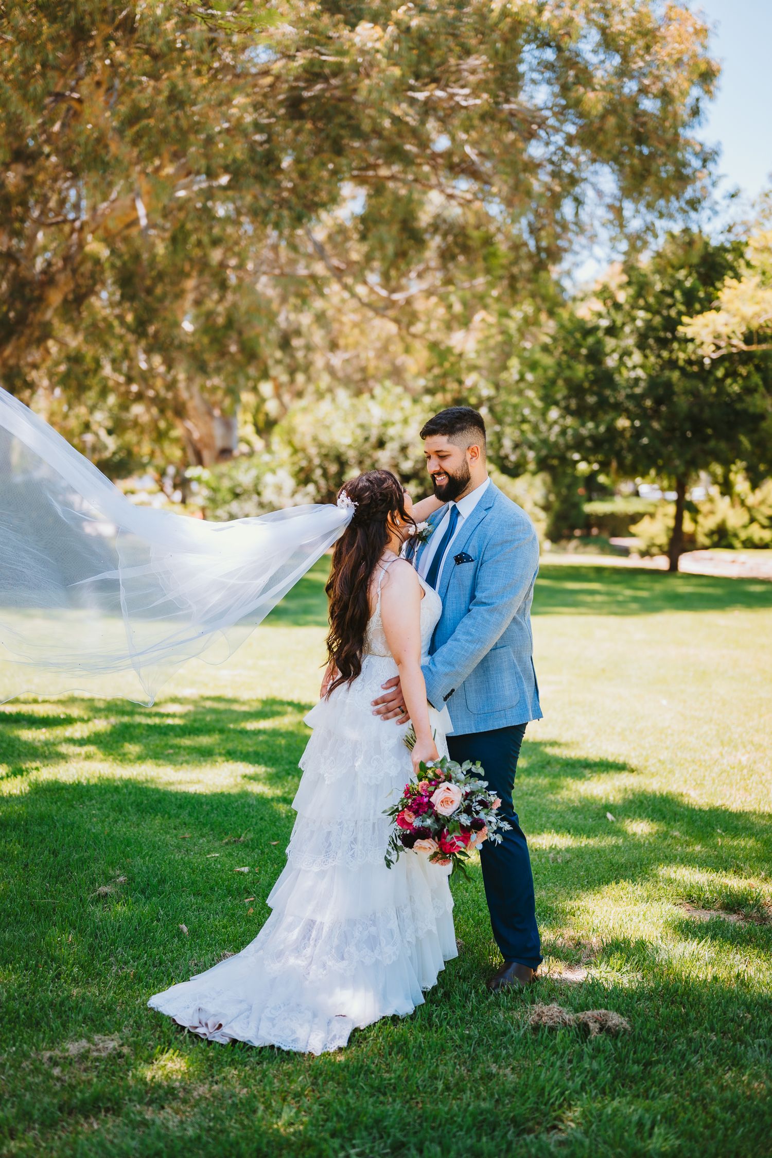 A newlywed couple embraces on a sunny lawn in front of trees while the bride's veil flows in the breeze.
