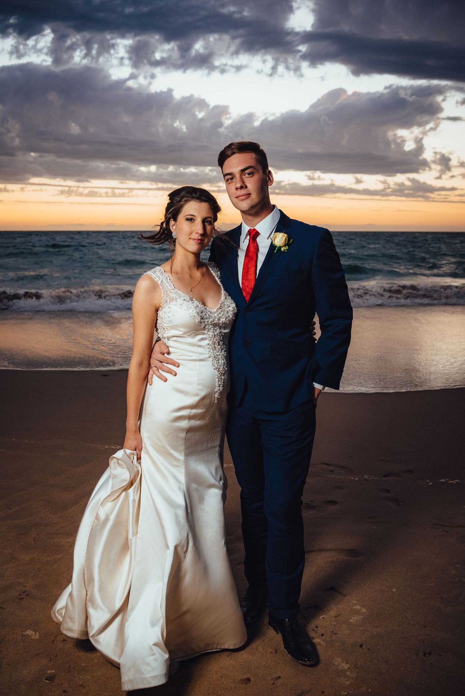 A wedding couple stands on the beach at sunset with dramatic clouds overhead and ocean waves in the background.