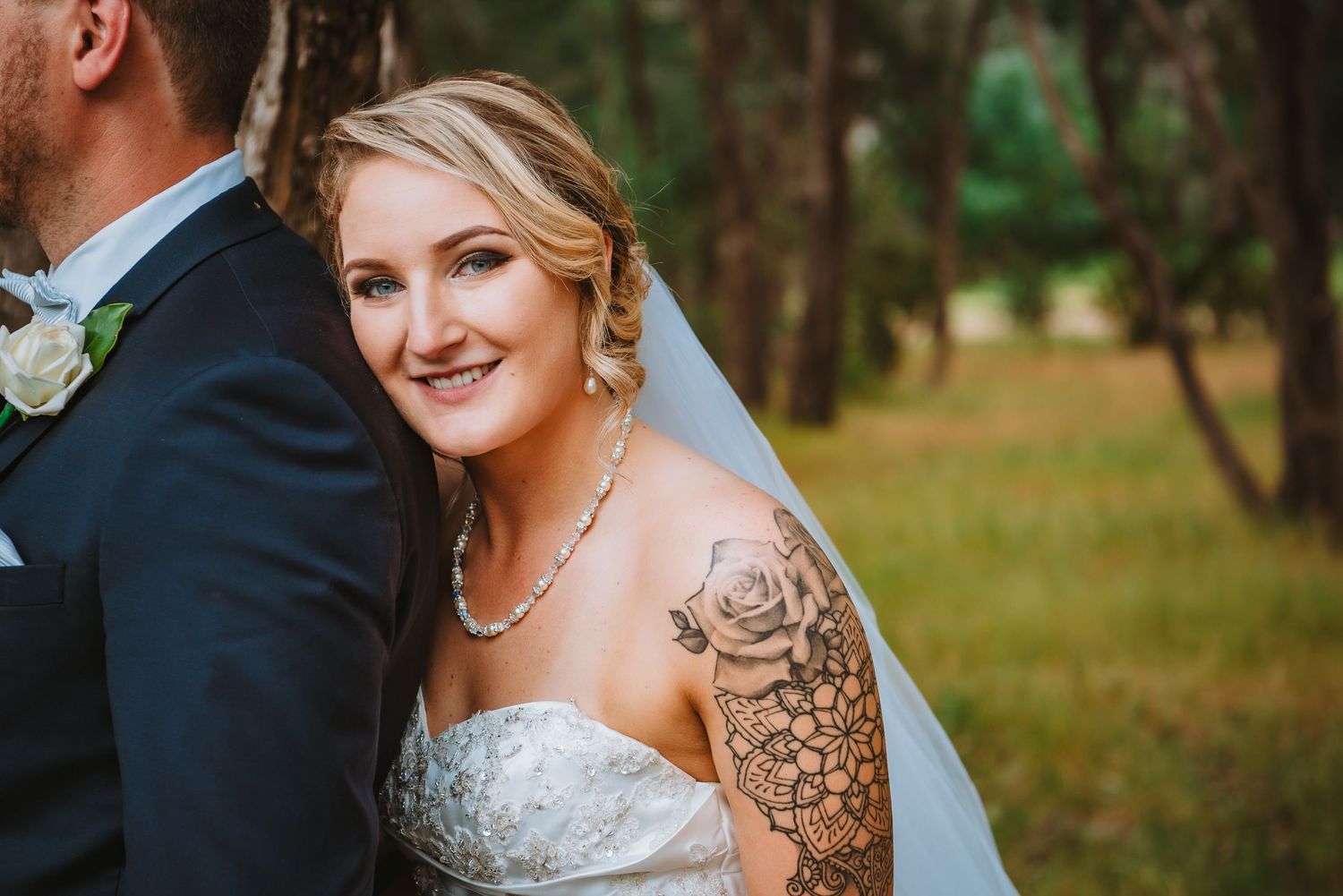 Bride in strapless beaded wedding dress with floral arm tattoo and veil smiles in outdoor woodland setting.