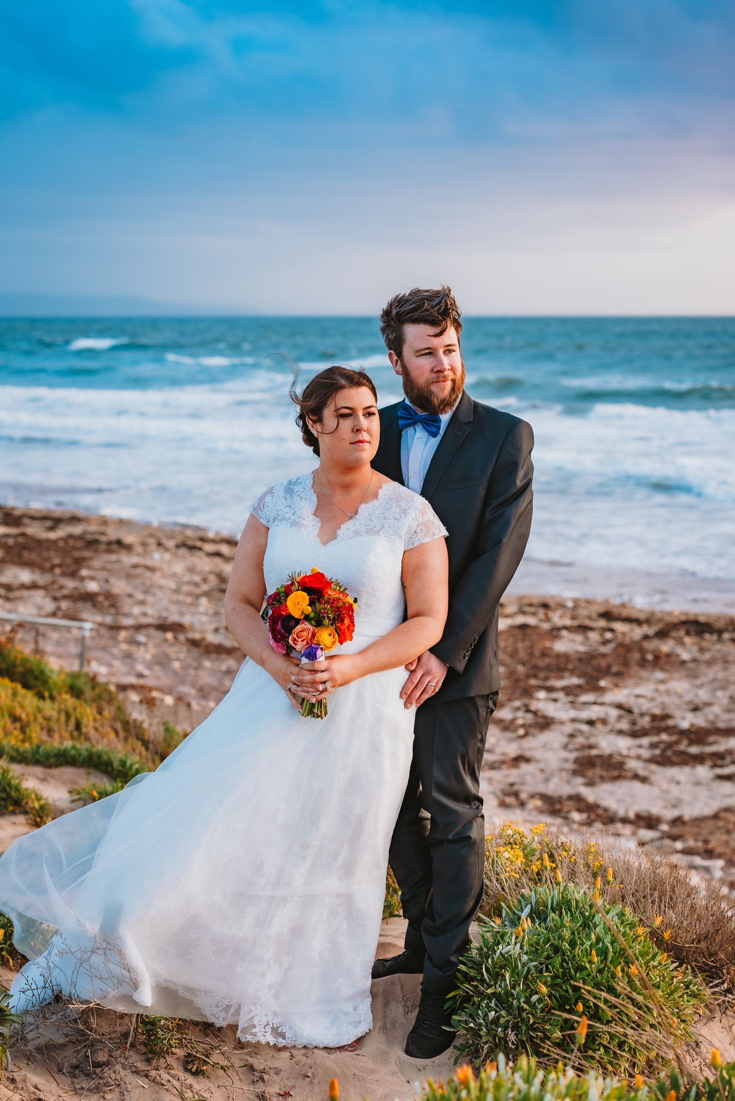 A couple in wedding attire stands together on a coastal cliff overlooking the ocean with the bride holding a colorful bouquet.