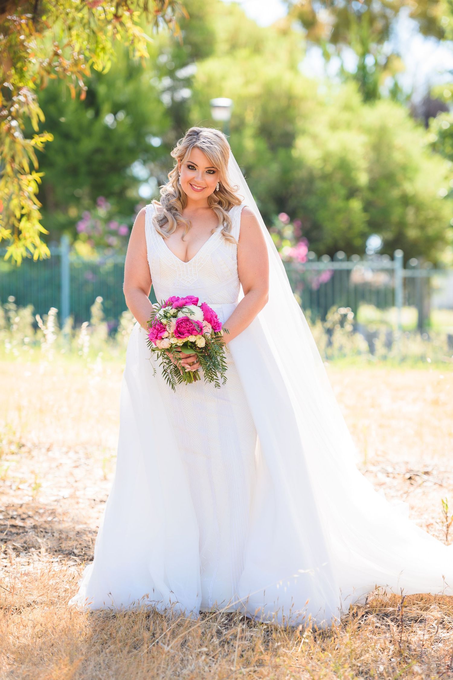 A bride in a flowing white lace wedding gown holds a bright pink bouquet while standing in a sunlit outdoor setting.