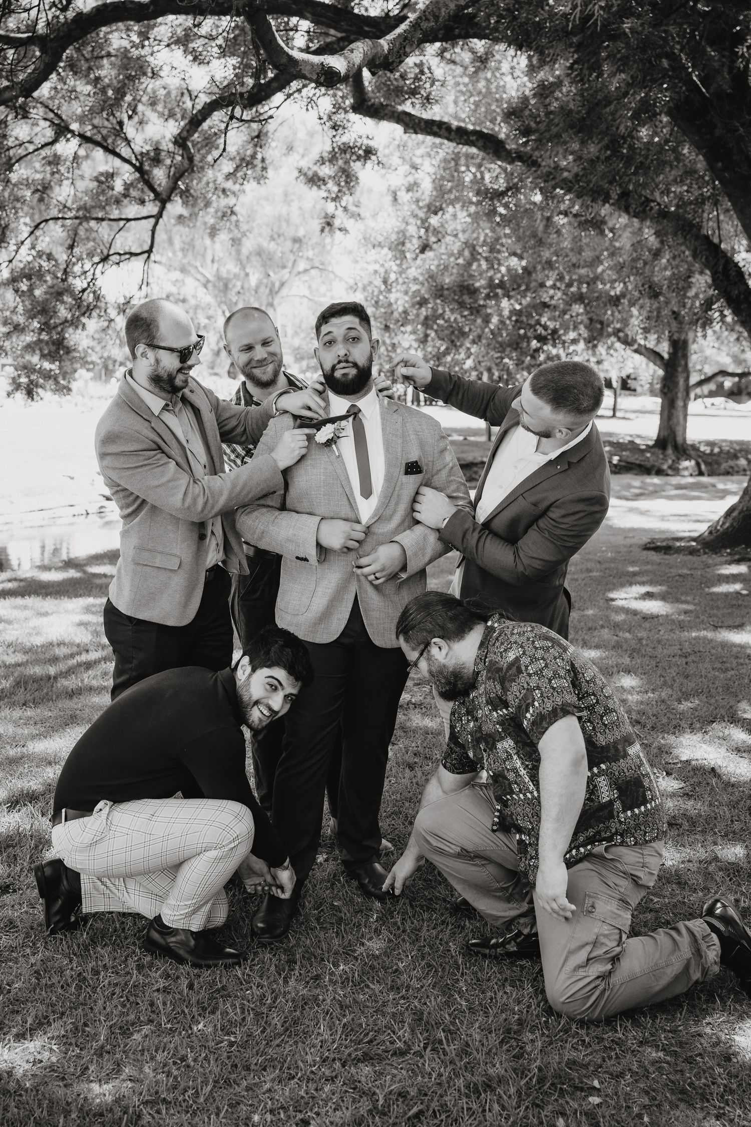 Playful black and white wedding party photo showing groomsmen adjusting the groom's bowtie outdoors under trees.