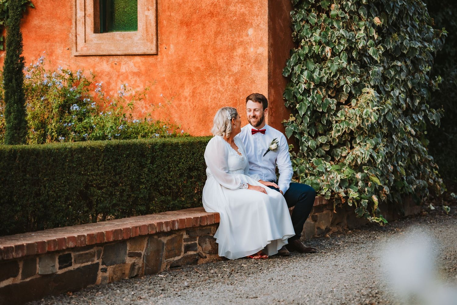 A couple sits together on a brick wall against a vibrant orange Mediterranean-style wall with greenery.