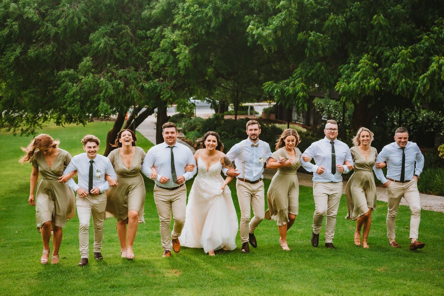 Wedding party in tan and blue attire jumps joyfully on green lawn during outdoor summer celebration.
