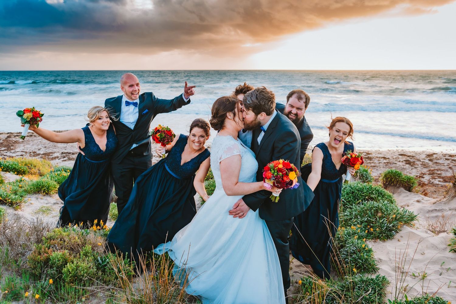 A wedding party in navy blue attire and white gown pose joyfully on a scenic coastal cliff at sunset.