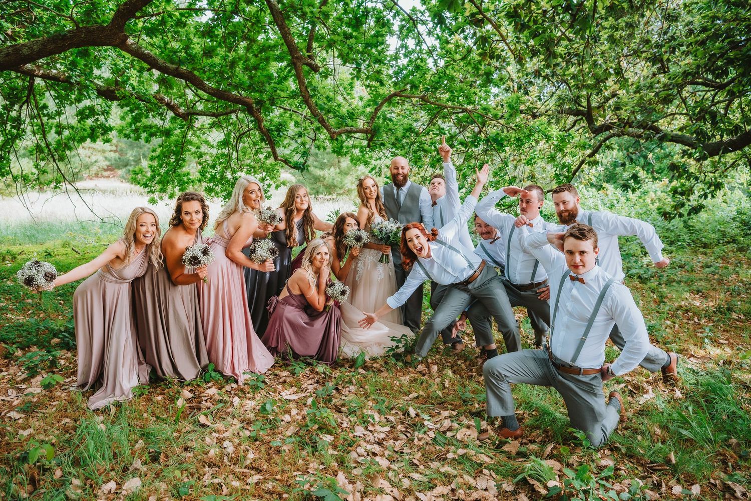 Wedding party poses playfully under green trees in a natural outdoor setting with bridesmaids in pink and groomsmen in gray.