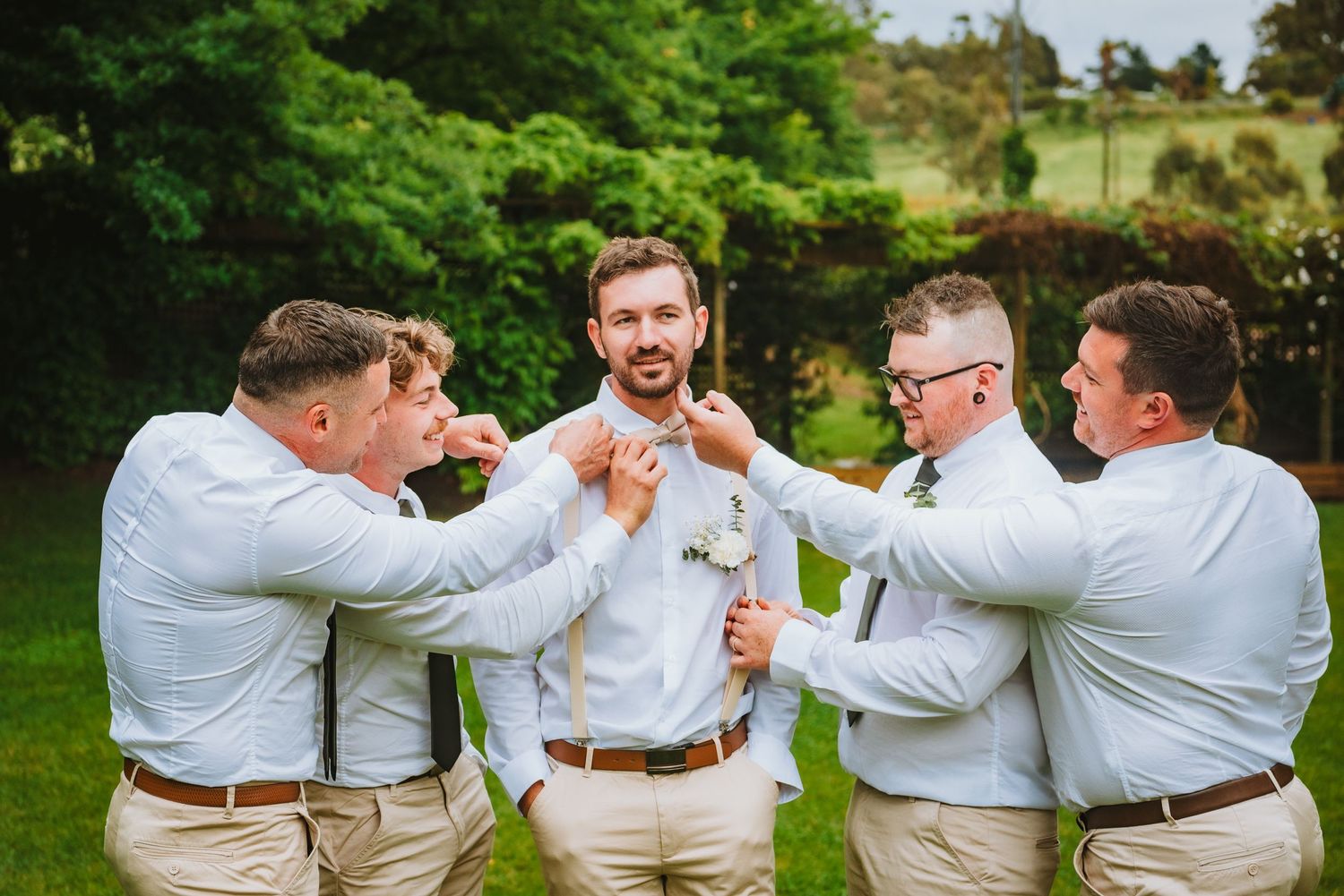 Groomsmen in matching light-colored attire and suspenders share celebratory champagne moment outdoors in garden setting.