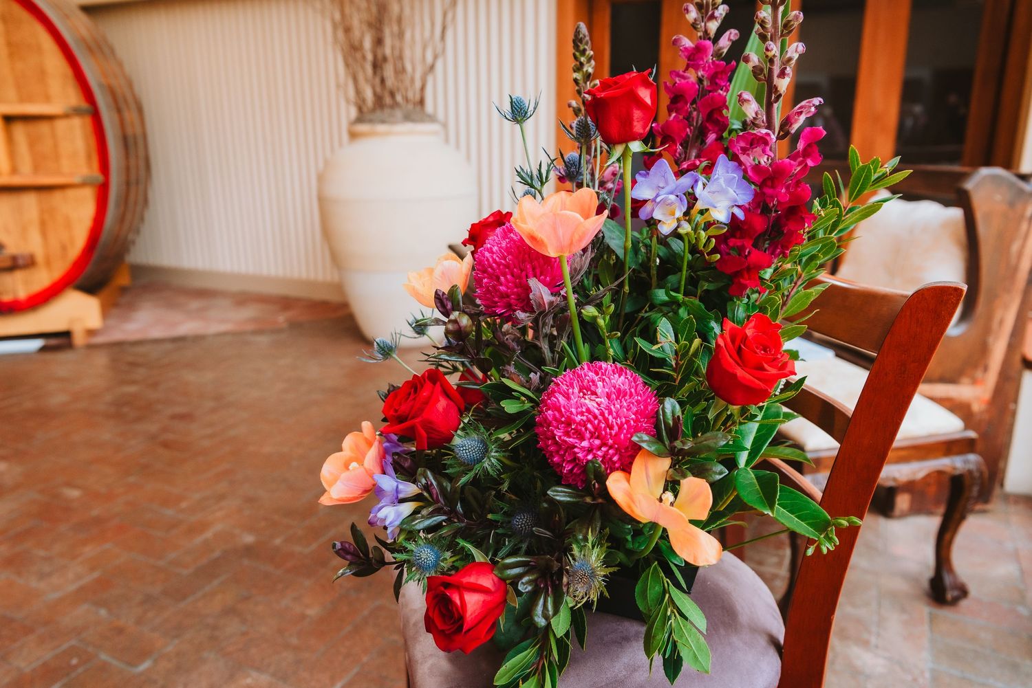A vibrant floral arrangement with pink dahlias, red roses and orange blooms sits in a vase on a rustic patio setting.