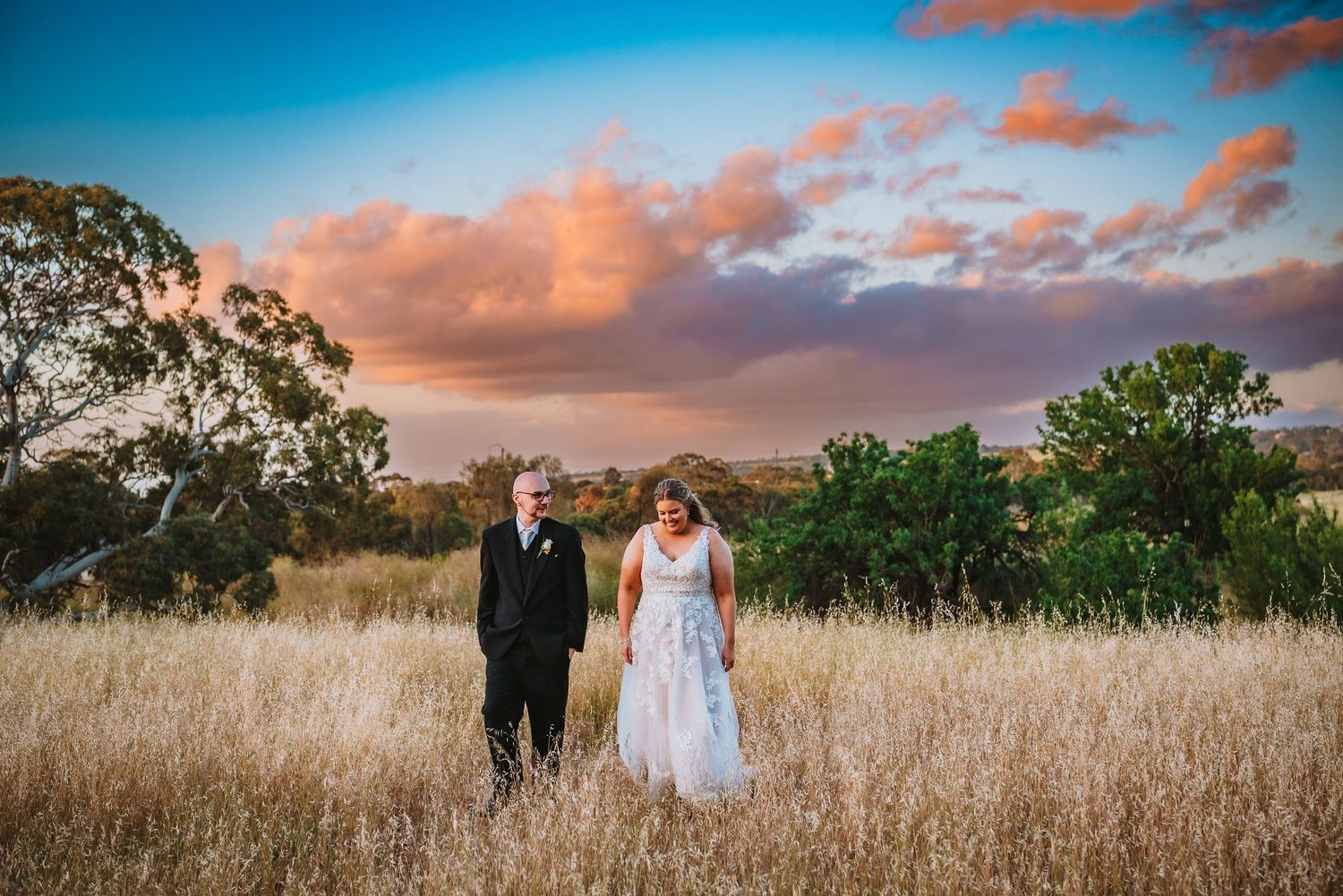 A couple stands in tall golden grass with dramatic sunset clouds and trees in the background.