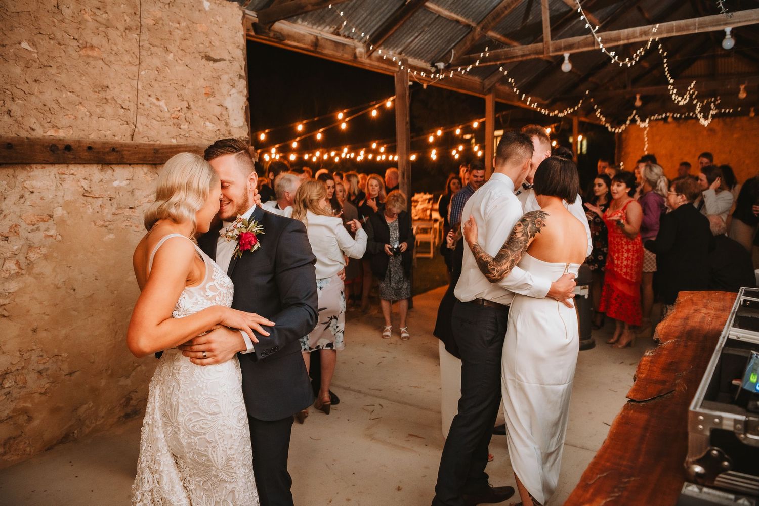 Couples slow dance together under warm string lights at a rustic venue during an indoor wedding reception.