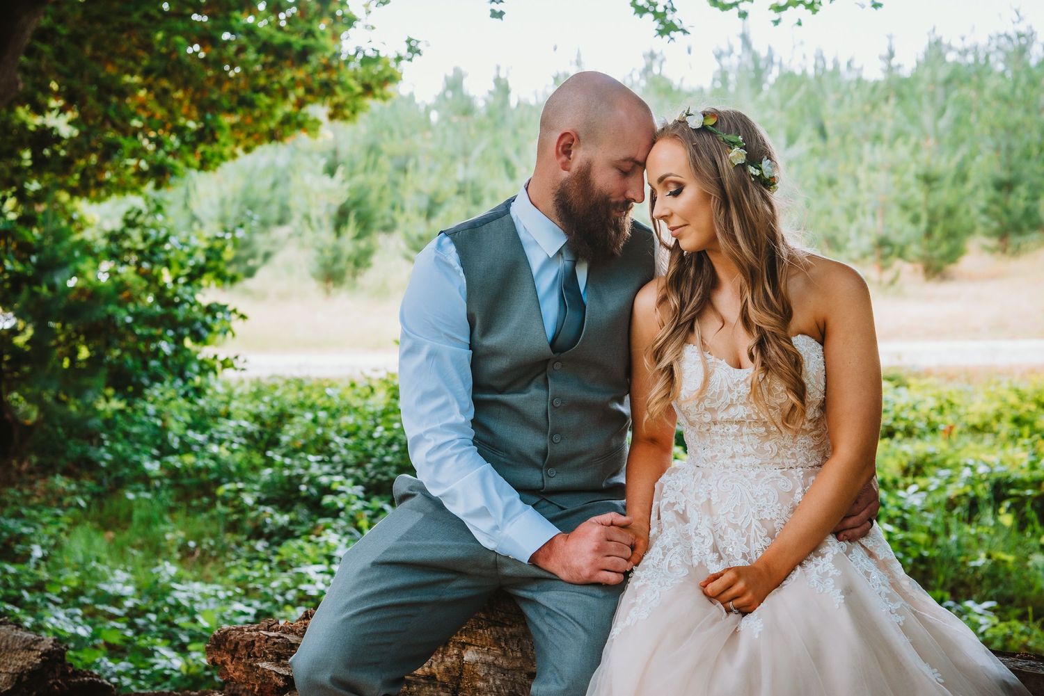 Newlywed couple sits intimately together outdoors surrounded by green foliage on their wedding day.