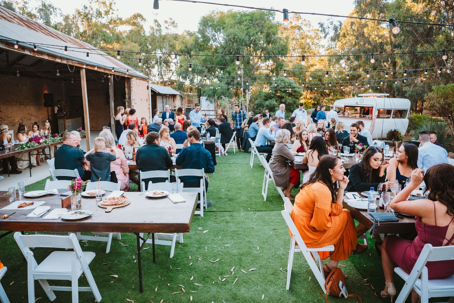 Outdoor wedding reception with guests seated at white tables on a lawn near rustic buildings at sunset.