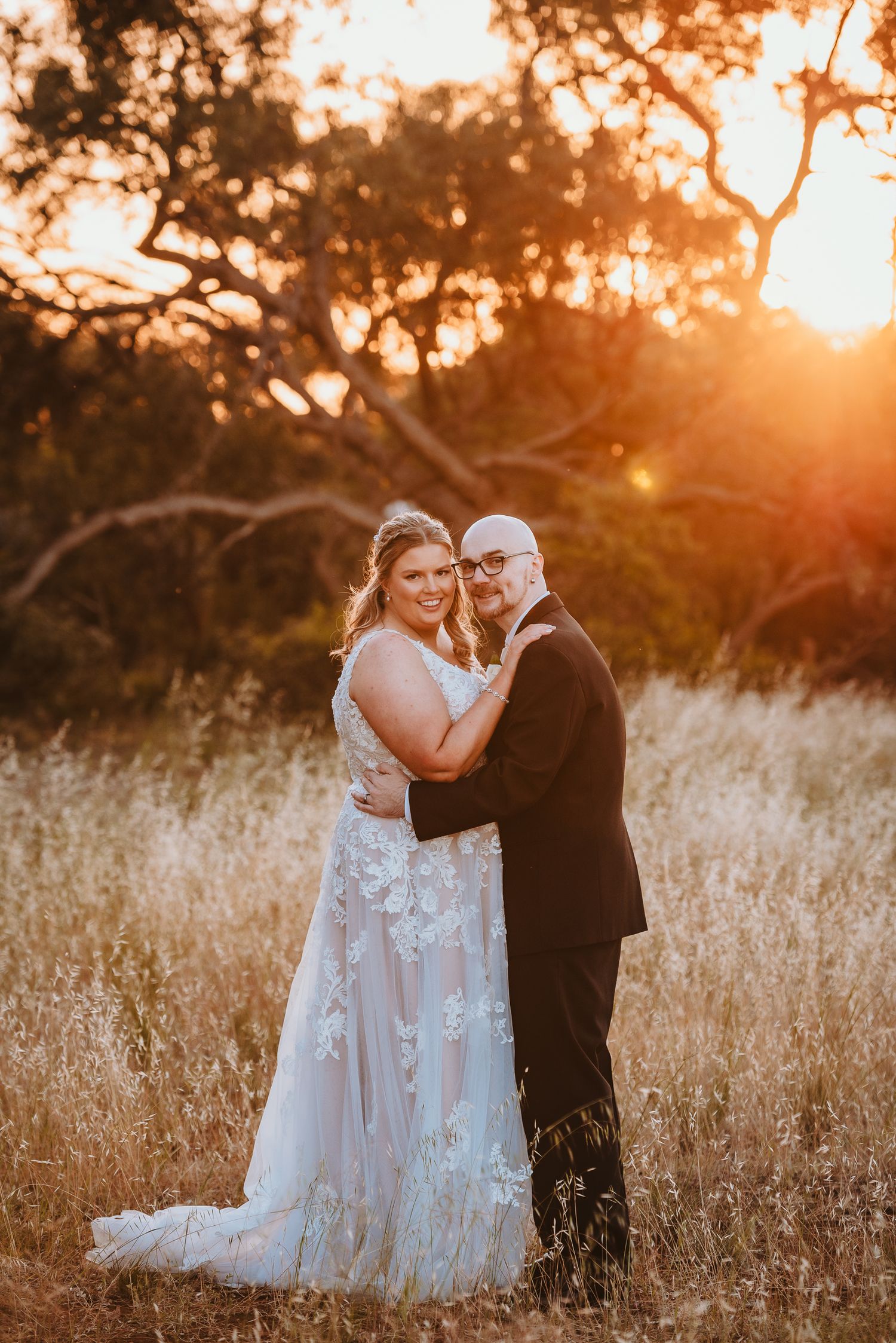 A romantic sunset wedding photo of a couple embracing in a golden field with trees silhouetted in the background.