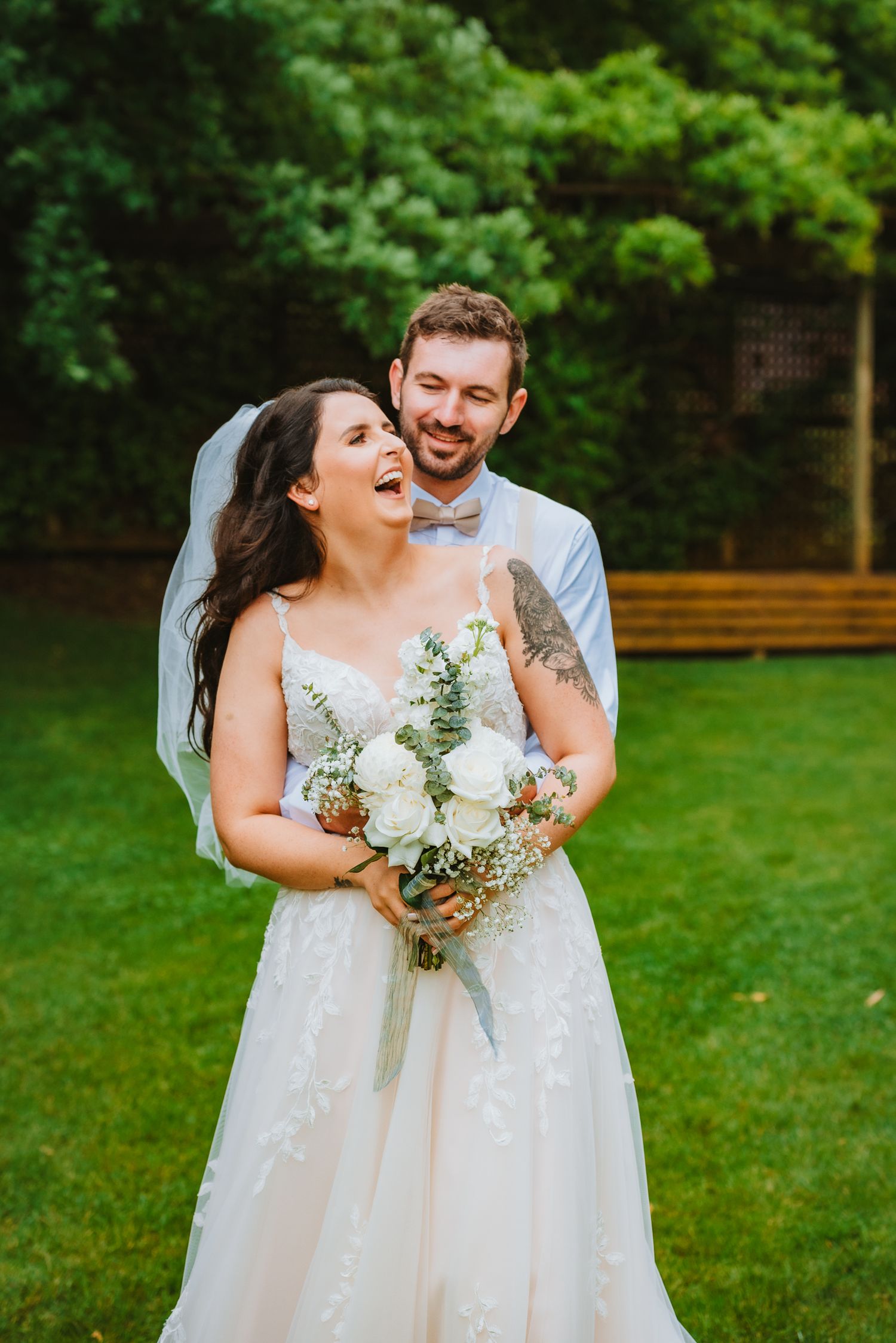 A joyful outdoor wedding scene with the couple laughing together while holding a white floral bouquet.