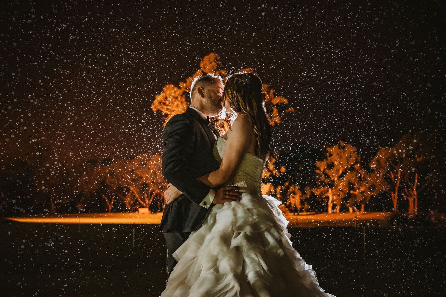 A couple shares a romantic moment under falling snow illuminated by warm amber lighting at night.