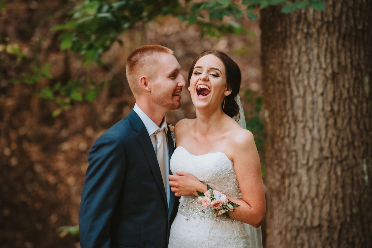 A joyful candid moment between newlyweds laughing together outdoors by a tree trunk.