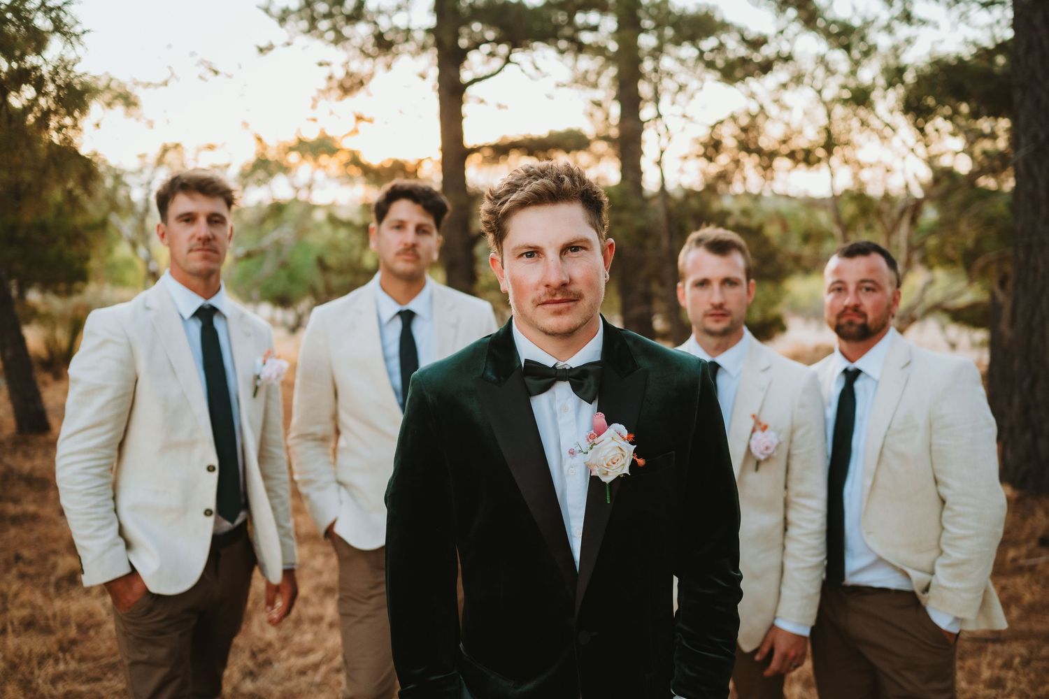Groom in black tuxedo stands in front with groomsmen wearing cream colored jackets behind at outdoor wedding venue.